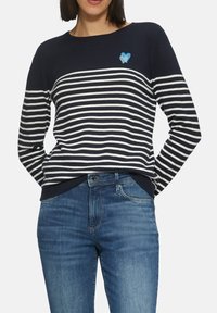 Gestreifter Pullover in Marineblau und Weiß mit rundem Ausschnitt, ausgestattet mit einem blauen Herz-Emblem. Kombiniert mit blauen Jeans.