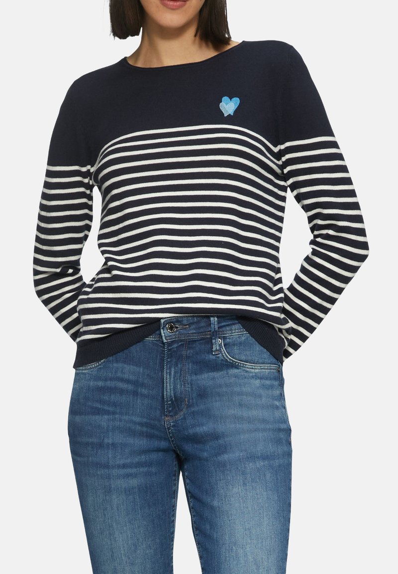 Gestreifter Pullover in Marineblau und Weiß mit rundem Ausschnitt, ausgestattet mit einem blauen Herz-Emblem. Kombiniert mit blauen Jeans.