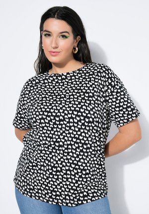 Donna con capelli lunghi e scuri che indossa una maglietta nera con motivi a cuoricini bianchi e jeans blu, posando davanti a uno sfondo bianco semplice.