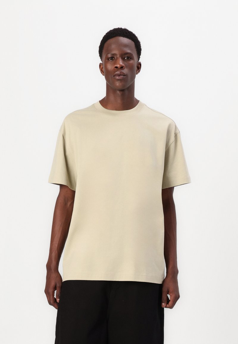 T-shirt en coton beige avec un col rond et des manches courtes, coupe décontractée, porté avec un bas noir, design simple, sans motifs ni embellissements.
