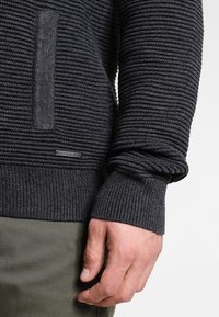Mörkgrå stickad tröja med ribbad textur, med en framficka och en logotypapplikation. Långa ärmar med manschettdetaljer och kontrasterande tyg.