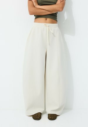 Personne portant un pantalon large blanc ample avec une taille à cordon et des chaussures vertes tissées à enfiler, debout les bras croisés.