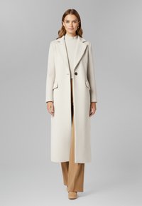 Donna che indossa un lungo cappotto crema testurizzato sopra un dolcevita crema e pantaloni ampi color cammello, in piedi davanti a uno sfondo grigio semplice.