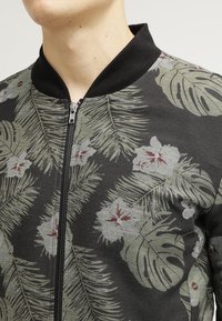 Uomo che indossa una giacca nera con zip, decorata con motivi di foglie tropicali verdi e fiori bianchi, con un colletto a coste nero.