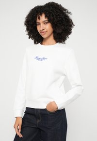 Witte sweatshirt met een ronde halslijn en geribbelde mouwen. Voorzien van blauwe geborduurde tekst "Guess Jeans" op de voorkant. Gemaakt in combinatie met donker denim broek.