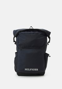 Zaino blu navy con design a rotolo, tasca frontale con zip e tasche laterali. Presenta il logo bianco "HILFIGER" alla base.