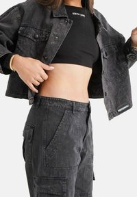 Persona che indossa un top a coste nero corto e una giacca di jeans grigio scuro con pantaloni cargo, sollevando leggermente la giacca per mostrare il ventre.