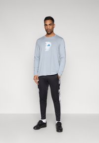 Light gray long-sleeve shirt with blue logo, paired with black tapered sweatpants featuring white side stripes and black sneakers.
