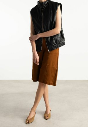 Personne portant un gilet en cuir noir sans manches avec une fermeture éclair, un short marron longueur genou et des talons ouverts couleur beige, debout sur un sol uni.