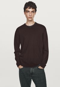 Dunkelbrauner V-Ausschnitt-Pullover mit langen Ärmeln, aus einem weichen Material gefertigt, über einem blauen Oberteil getragen, kombiniert mit blauen Jeans. Einfaches Design, ohne Muster.