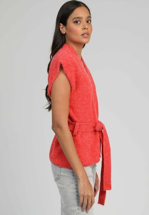 Jeune femme aux longs cheveux portant un gilet en maille corail sans manches noué à la taille et un jean gris clair à studs sur le côté, vue de profil.