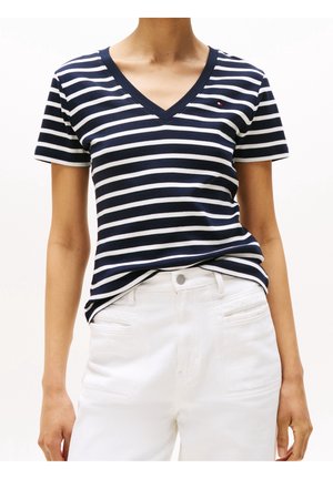 Vrouw draagt een marineblauw-wit gestreept V-hals T-shirt, ingestopt in een witte broek met hoge taille, voorzakken en knoopsluiting.