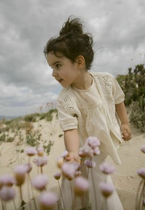 Blusa color crema con cuciture decorative, abbinata a pantaloni di lino leggeri. Bambino che si allunga verso piccoli fiori rosa in un contesto sabbioso.