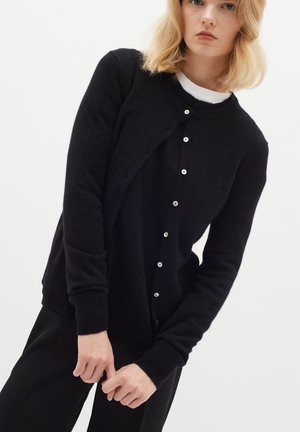 Gilet noir en tricot fabriqué dans un tissu doux, avec un col rond et une fermeture à boutons avec de petits boutons blancs ronds. Manches longues.