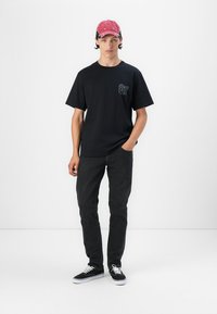 Camiseta negra de manga corta con un logo en el pecho, combinada con jeans oscuros y zapatillas negras. El modelo lleva una gorra rosa. Fondo minimalista.