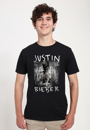 Jeune homme souriant aux cheveux bouclés portant un t-shirt noir Justin Bieber et un pantalon beige sur un fond blanc uni.