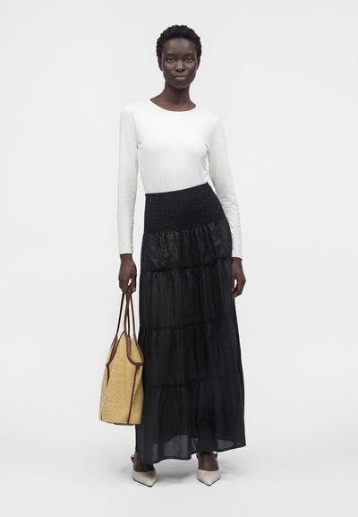 Femme portant un haut blanc à manches longues et une jupe longue noire à volants, tenant un sac fourre-tout beige avec une bordure marron, debout devant un fond uni.