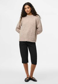 Mlada ženska z dolgimi kodrastimi lasmi, oblečena v bež puhasto pulover, črne kratke hlače in črne balerinke, ki stoji z rokami za hrbtom.