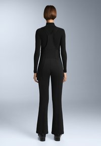 Černý overal s dlouhými rukávy, vysokým výstřihem a rozšířenými nohavicemi; má strukturovanou tkaninu a zipy na kotnících pro nastavitelný střih.