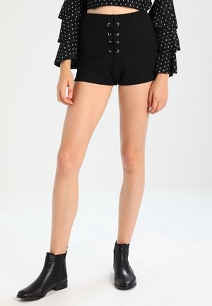 Frau trägt schwarze Schnürshorts, schwarze Stiefeletten und ein schwarzes Langarmshirt mit weißen Tupfen und stufigen Ärmeln.