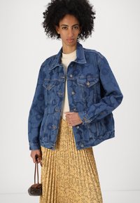 Jeans jakna v temno modri barvi s cvetličnim vzorcem, s klasičnimi gumbi, dvema žepoma na prsih in sproščenim krojem, oblečena čez bež top.