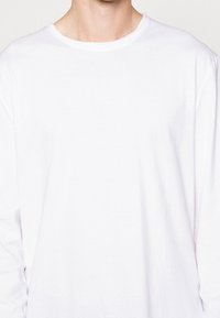 Camisa blanca de manga larga hecha de algodón ligero. Presenta un cuello redondo y un corte relajado, con una textura suave y sin patrones visibles.