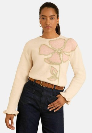 Cremfarbener Pullover mit texturierten Blumenstickereien in Pink und Grün, mit langen Ärmeln und einem gerippten Kragen. Kombiniert mit dunklen Jeans.