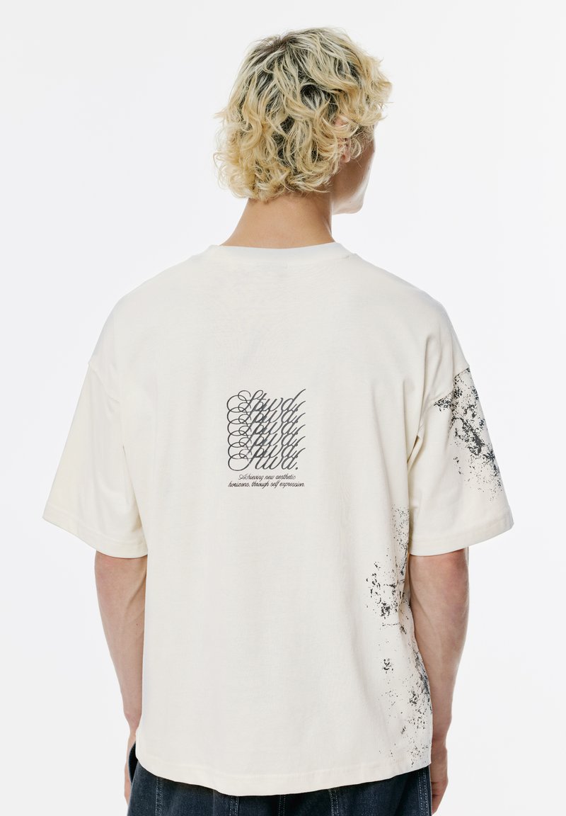 Tricou din bumbac crem cu croială lejeră, având text grafic negru pe spate și detalii cu stropi de cerneală pe mânecă și pe lateral.