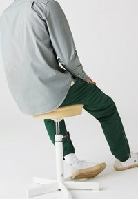 Chemise à motif gris, pantalon vert, assis sur un tabouret crème avec une base blanche. Le tabouret dispose d'une fonction de hauteur réglable.