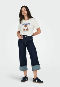 Grafisk t-shirt i creme med en bjørneillustration, parret med mørk denim bukser med brede ben og opslåede manchetter samt navy sneakers.