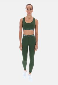 Olivovo zelený športový crop top a legíny s bielym logom na páse, spárované s bielymi športovými topánkami. Hladká textúra.