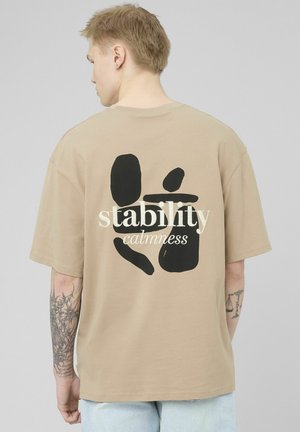 Personne blonde avec des tatouages portant un T-shirt beige avec des formes abstraites noires et les mots « stabilité sérénité » au dos, fond clair.
