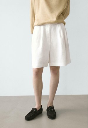 Persoon die losse, witte korte broek tot net over de knie draagt, een beige top met lange mouwen en zwarte veterschoenen, staand op een grijze vloer.