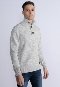 Maglione grigio a maglia con collo alto, dotato di tre bottoni e polsini a coste. La trama è variegata con sottili motivi sparsi.