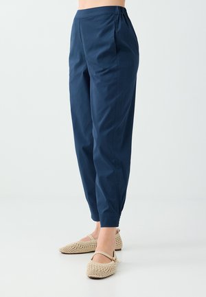 Paire de pantalons bleu marine fuselés portés avec des ballerines tissées beige Mary Jane sur un fond clair uni.