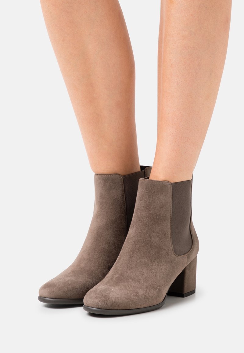 Bottines basses en daim taupe avec talons blocs portées sur jambes nues sur un fond clair uni.