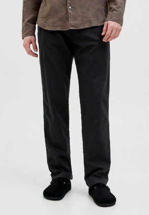 Homme debout portant un pantalon noir à jambes droites, des chaussures noires sans lacets, et une chemise marron à boutons, sur un fond blanc uni.