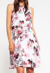 Robe fleurie avec un design sans manches, en tissu léger, et des motifs de fleurs roses et violettes sur fond blanc.