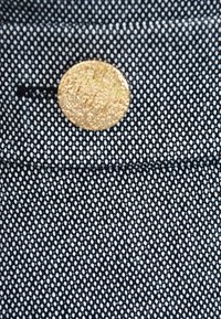 Czarna i biała teksturowana tkanina z wzorem w kratkę, z okrągłym złotym guzikiem przyszytym na górze. Guzik ma szczotkowane metaliczne wykończenie.