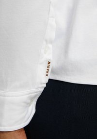 Biała bawełniana koszula z zapięciem na guzik na rękawach, z metką oznaczoną napisem "CHASIN'" w brązowym kolorze, wyeksponowana na ciemnym tle.