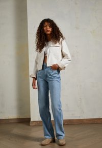 Giacca di jeans bianca cropped, jeans blu a vita alta e stivali alla caviglia in suede beige, su uno sfondo neutro chiaro con pavimento in legno.