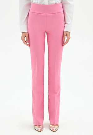 Pantaloni rosa dritti con vita alta indossati con una camicia bianca a maniche lunghe e tacchi metallici con punta a mandorla, in piedi di fronte.