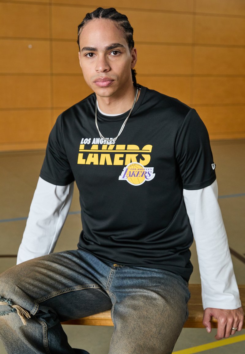 Črno atletsko majico s logotipom "Los Angeles Lakers", kratkimi rokavi, oblečeno čez belo majico z dolgimi rokavi. V paru z raztrganimi modrimi kavbojkami.