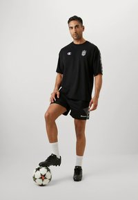 Juoda sportinė marškinėliai su logotipu, derinami su juodais šortais. Modelis stovi ant futbolo kamuolio, dėvi juodus sportinius batelius ir baltas ilgas kojines.