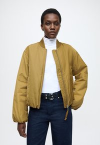 Musztardowo-żółta kurtka bomberka z zapięciem na zamek błyskawiczny, ściągaczem przy kołnierzu i elastycznymi mankietami. Połączona z białym golfem i ciemnymi dżinsami.