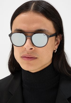 Uomo con lunghi capelli scuri che indossa occhiali da sole rotondi riflettenti, un dolcevita nero, un blazer nero e un piccolo orecchino a cerchio.