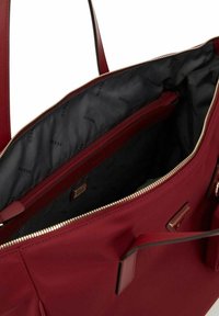 Bolso tote rojo con un exterior suave, cierre de cremallera dorado y un interior negro con patrones de logo y un bolsillo con cremallera.