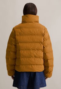Bruine pufferjack met hoge kraag, horizontaal doorgestikt ontwerp en een gladde textuur, gecombineerd met een denim rok.