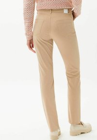 Pantalon beige ajusté en tissu élastique ; présente deux poches arrière et une petite étiquette sur la ceinture ; texture lisse.