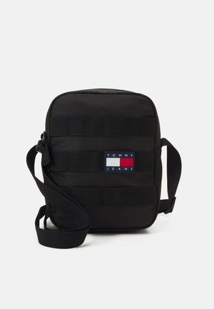 Borsa a tracolla nera con cinturini orizzontali in tessuto, etichetta con logo in rosso, bianco e blu. Presenta una chiusura con zip e una tracolla regolabile.